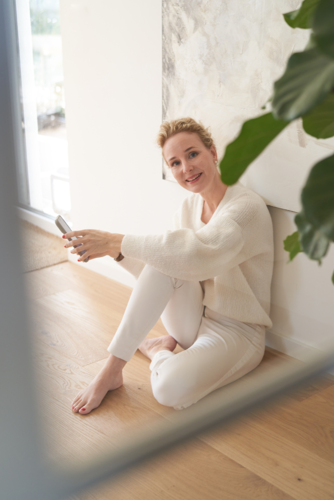 Frau sitzt mit Handy am Boden und schaut in die Kamera.